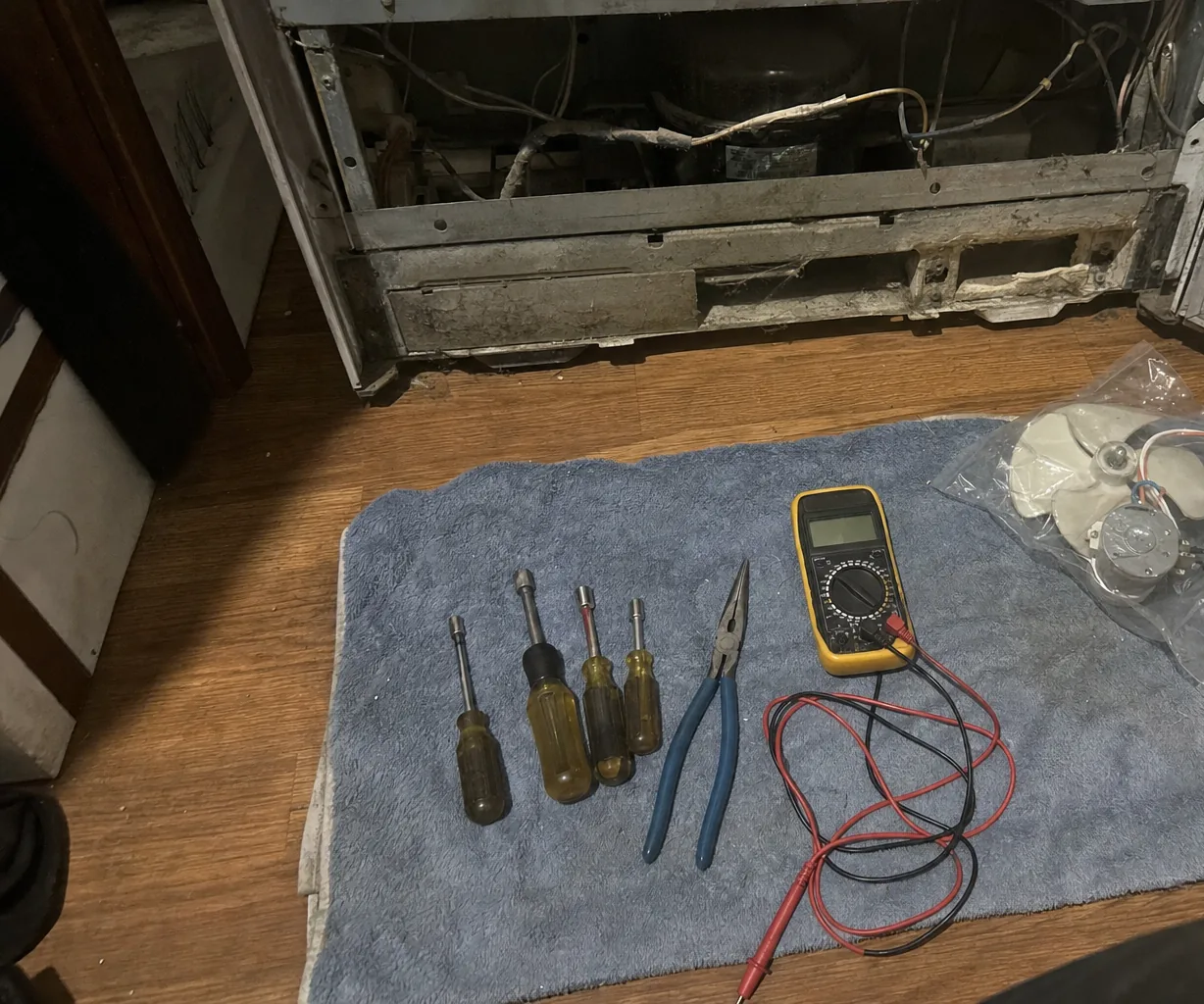 Kick plate off a refrigerator showing the dusty compressor bay and wiring, with nut drivers, blue pliers, a yellow multimeter and a bagged replacement fan laid on a towel - Gaggenau vs Sub-Zero Refrigerator