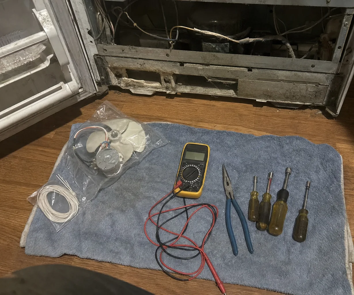 Refrigerator with the door open and the lower front panel removed to expose the compressor and wiring, with a multimeter, pliers, nut drivers and a bagged fan motor laid out on a blue towel