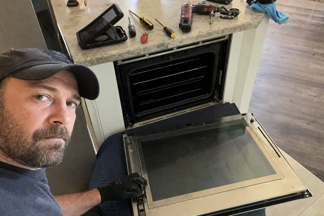 A wall oven door off its hinges on a moving blanket, hinge mechanism exposed, tools on the counter in frame - Appliance Repair in Nassau County